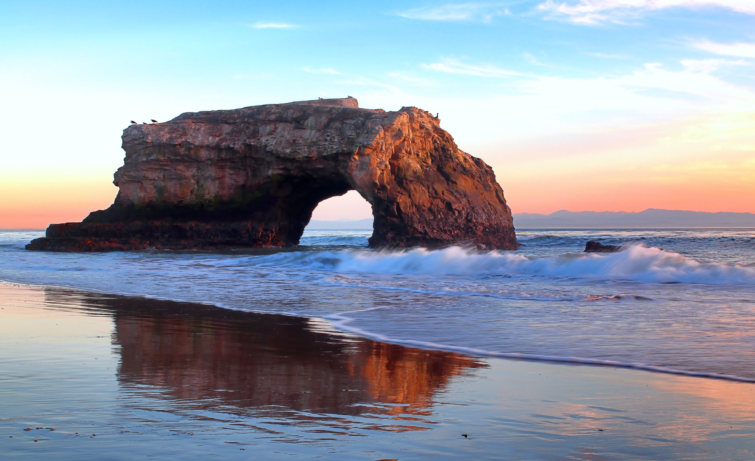 Natural Bridges State Beach (Santa Cruz) at sunset. In the medium distance, it shows a large, naturally occurring mudstone bridge carved by the Pacific Ocean into a cliff jutting out into the sea. A small wave is breaking as it approaches the sandy beach, which reflects the sunset in a thin layer of water. Photo courtesy of Simon Hurry, published on January 29, 2022, free to use under the Unsplash License.